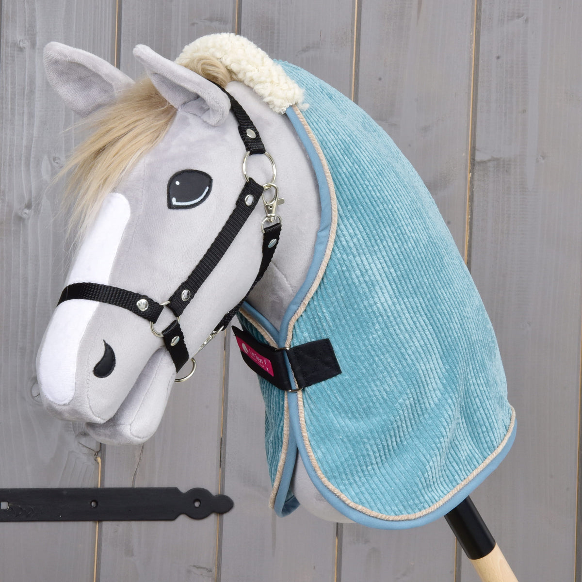 A plush hobby horse by Hobby Horse LarDen features a light gray face, black bridle, white muzzle, and wears the Stable Blanket Fern in light blue PES material, posed against a wooden background.