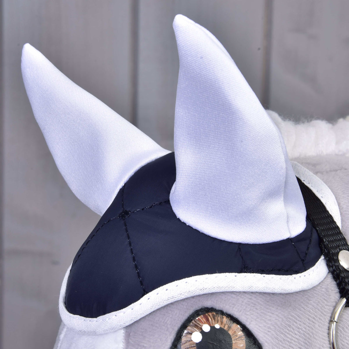 Close-up of a plush horse's head wearing the elegant Set Thermo Blue by Hobby Horse LarDen, featuring white ears, a navy and white cap, and black bridle. The perfect decorative piece against light wooden panels.