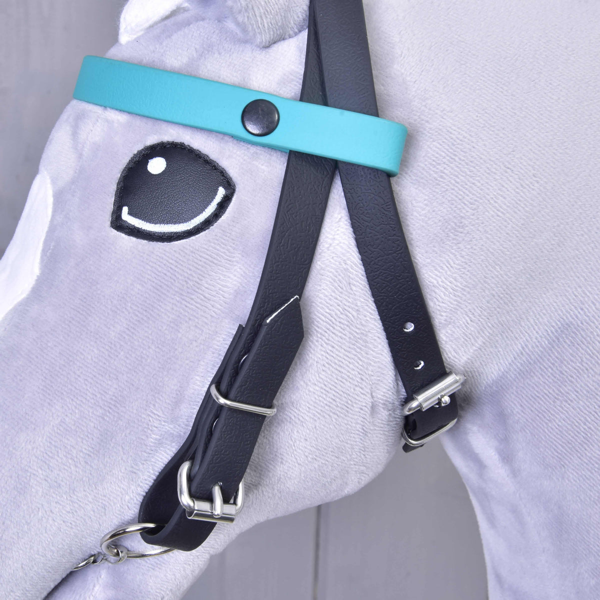 Close-up of the plain turquoise browband with a black press stud on the black bridle of a grey Hobby Horse