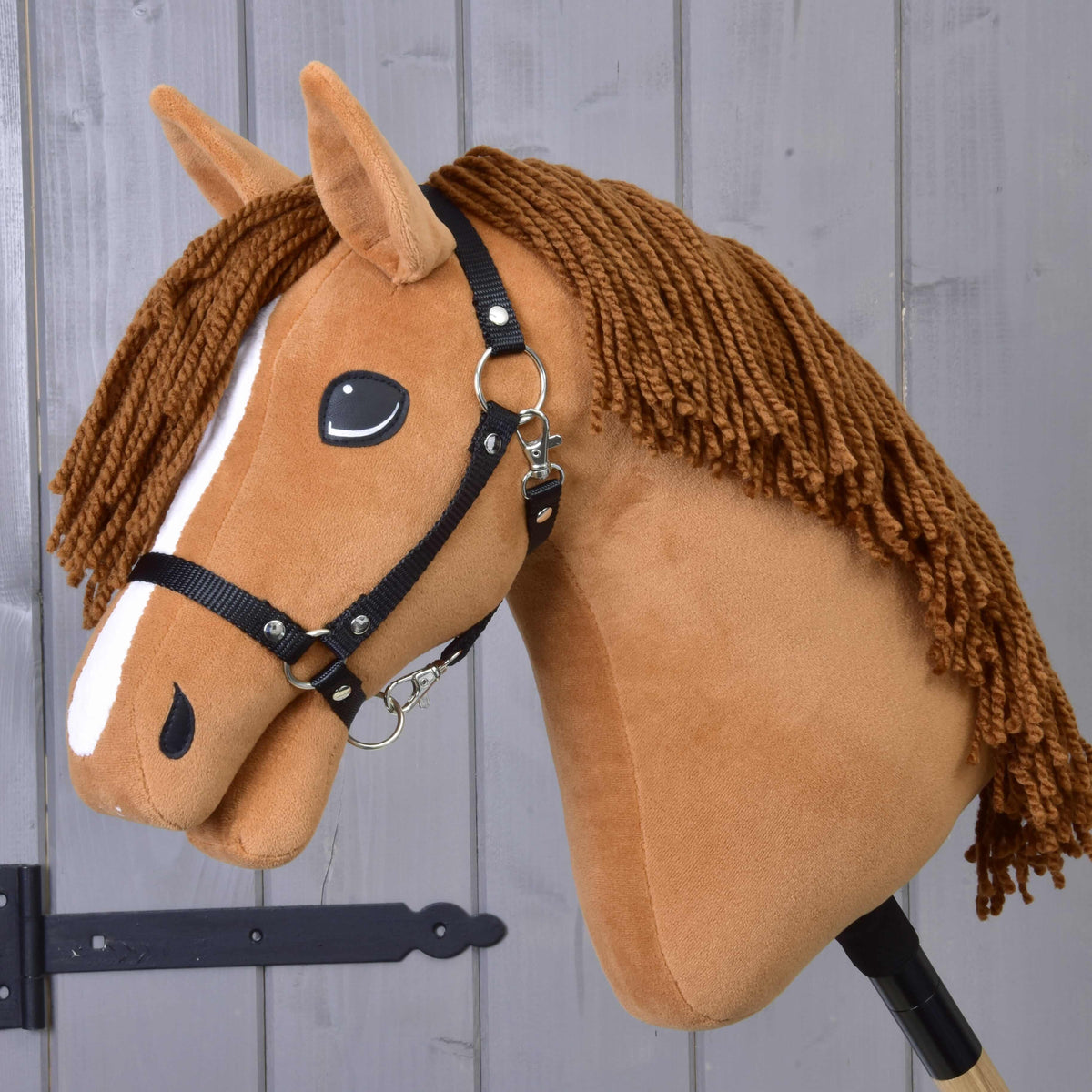 The Hobby Horse LarDen "Hobby Horse Wendy with Black Halter" is a chestnut mare plush toy featuring a brown yarn mane, white face stripe, and black halter—ideal for imaginative play—shown against a wooden panel background.