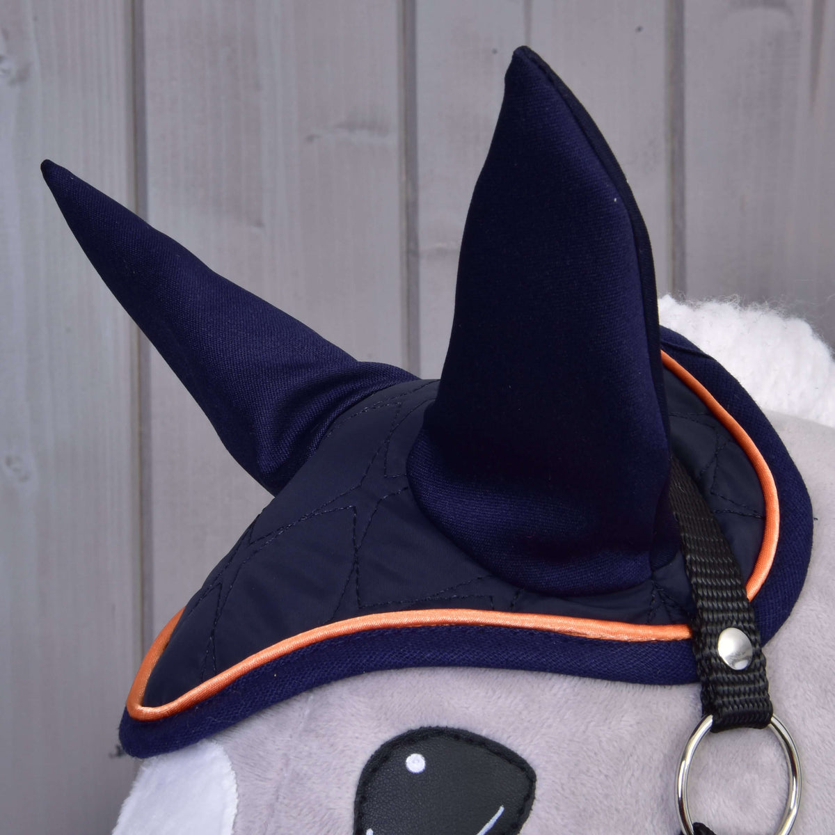 A close-up of the Set Winner by Hobby Horse LarDen shows a plush animal in a dark blue ear bonnet with pointed ears and orange trim, set against light gray wooden panels for a charming horse-themed decor look.