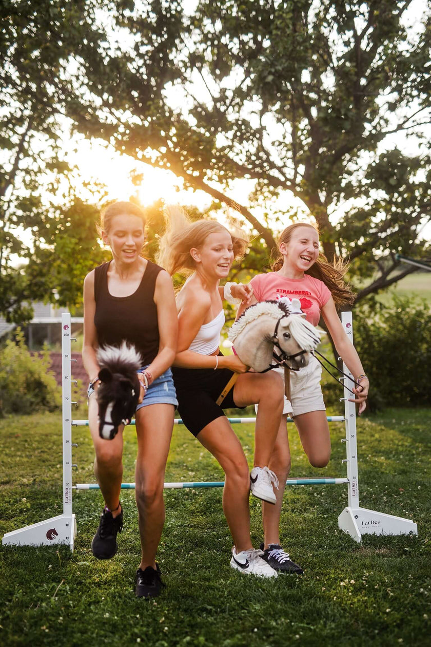 Three young women laugh as they run and jump over a hurdle outdoors, each holding a Hobby Horse, sunlight shining through the trees behind them as they enjoy the fun of Hobby Horsing.