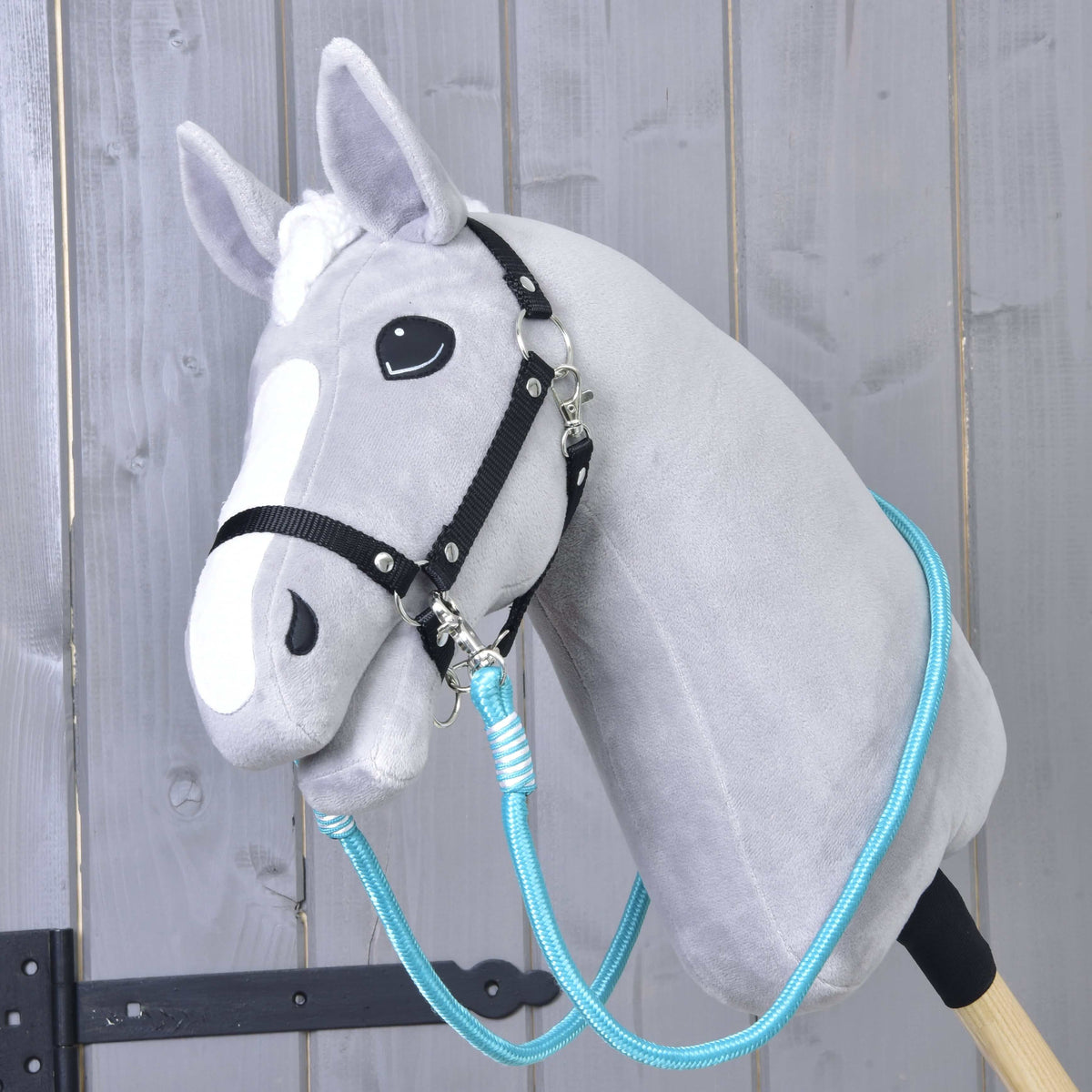 The Hobby Horse LarDen Reins Atlantis is a gray plush hobby horse with a black bridle and blue polypropylene rope reins on a wooden stick, shown in front of a rustic wooden wall.