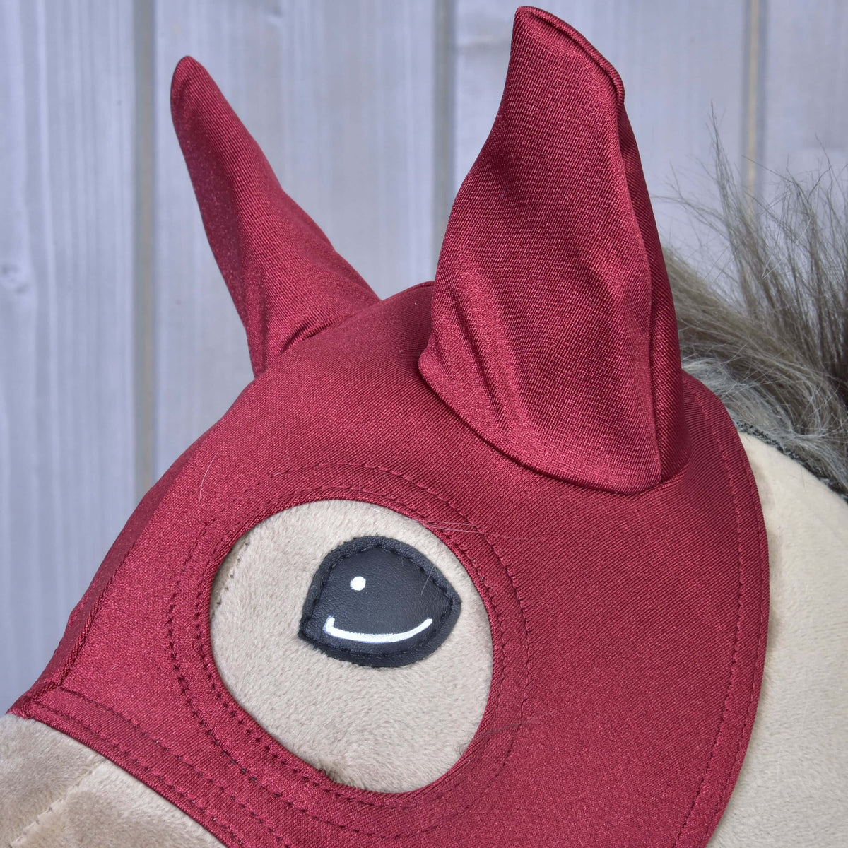 Close-up of a Hobby Horse LarDen plush toy featuring the Horse Mask Barn Red, with pointy ears and a grey mane, set against a light-colored wooden background.
