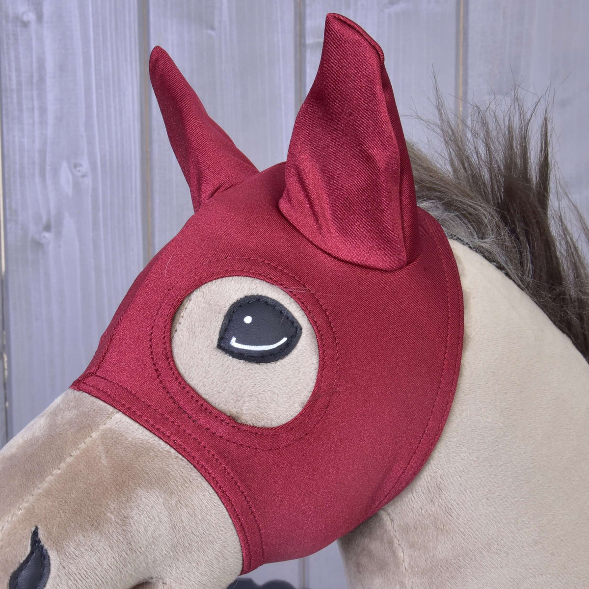 A beige plush hobby horse by Hobby Horse LarDen wears the Horse Mask Barn Red, featuring pointed ears and large cartoon-like eye openings, set against a wooden panel background.