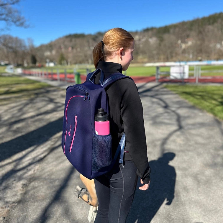 En ung kvinde med rødt hår i hestehale går udenfor i solskin i sort sportstøj med en marineblå rygsæk og en lyserød drikkedunk.