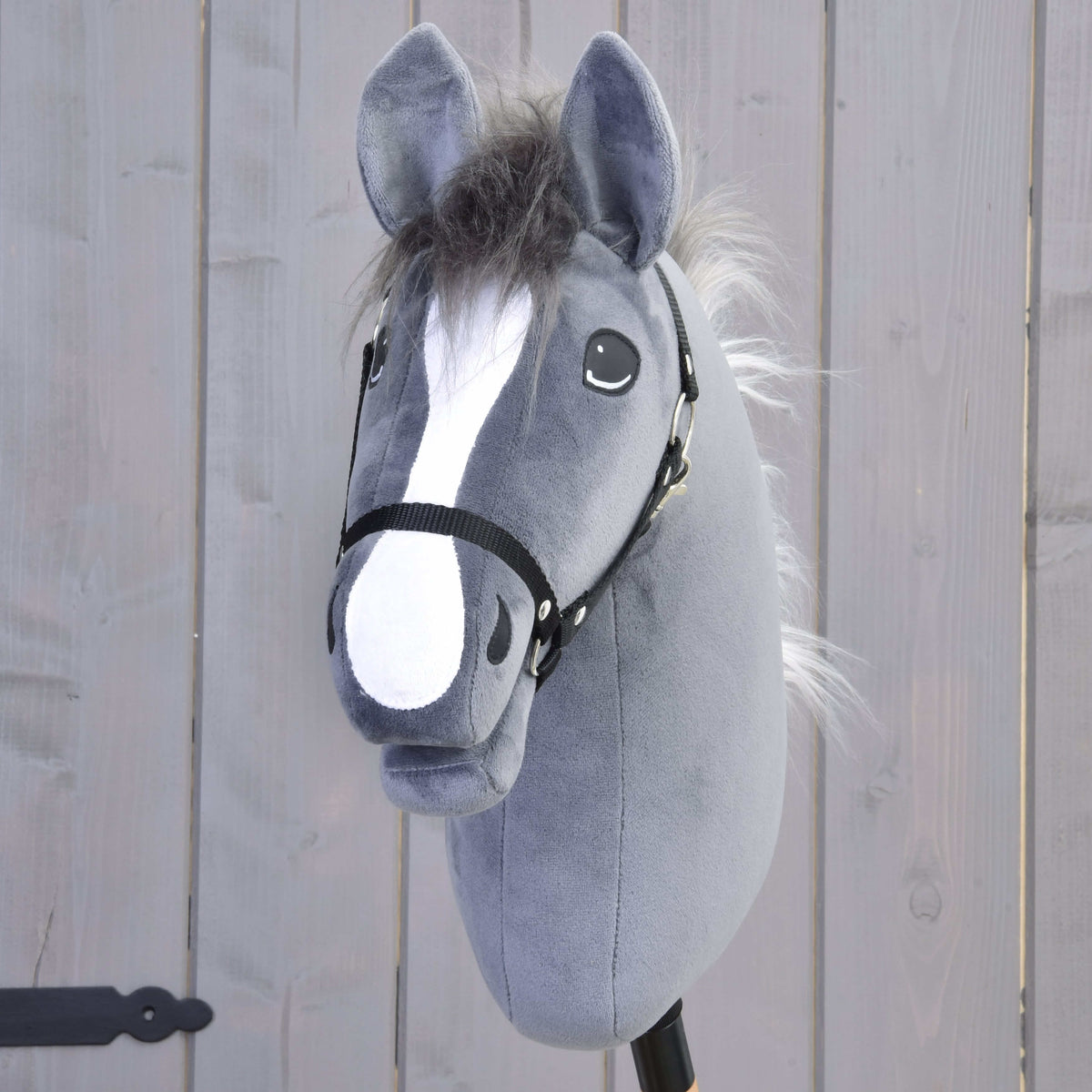 The Hobby Horse Beauty with Black Halter by Hobby Horse LarDen is a gray plush hobby horse with a black bridle, white blaze, and fluffy mane, inspired by an English Thoroughbred stallion and set against a light wood background.