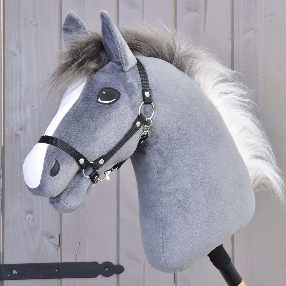 The Hobby Horse LarDen Hobby Horse Beauty with Black Halter features a gray plush stallion head with a white mane on a wooden stick, shown against a wooden fence background.