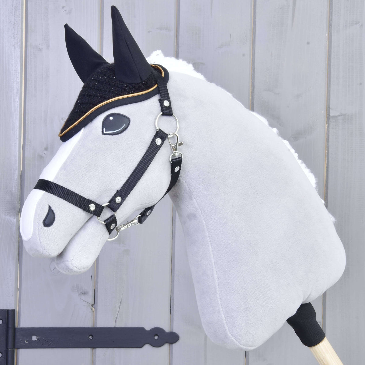 A gray Hobby Horse LarDen with black reins and a white mane wears the Ear bonnet crocheted Black Gold for insect protection, featuring black facial details, set against a light wooden background.