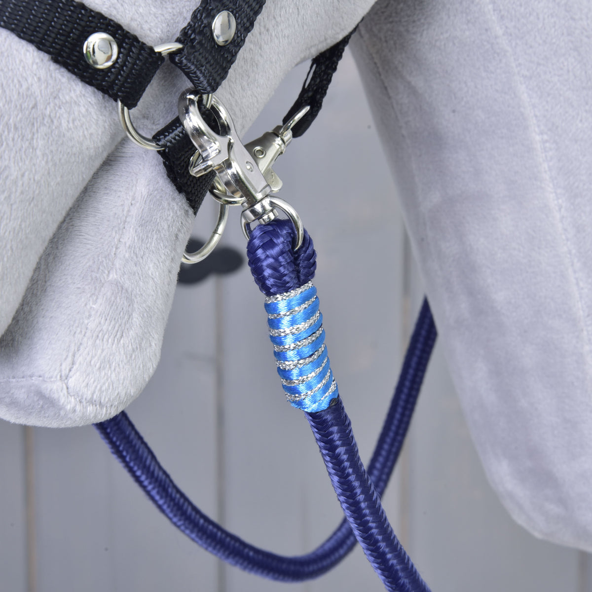 A close-up of the Hobby Horse LarDen Reins Blues, a blue and silver braided polypropylene lead rope, attached to a black halter on a grey horse against a wooden background.