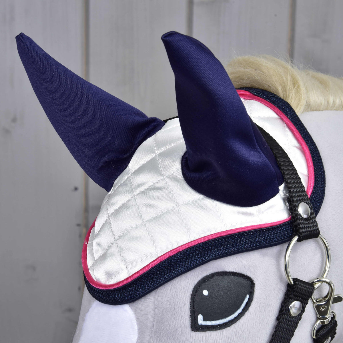 A close-up of a Hobby Horse LarDen's head features the Ear Bonnet Number 1—white quilted with dark blue ears and pink trim—for insect protection, plus a black bridle, against a light wooden background.