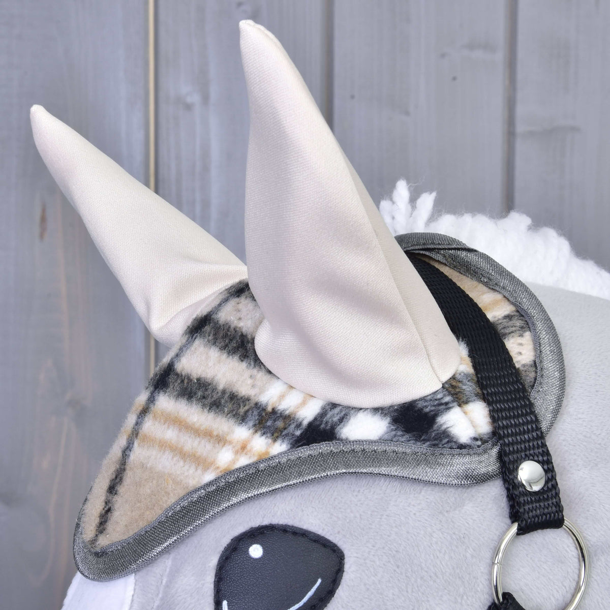Close-up of a Hobby Horse LarDen plush hobby horse with two beige fabric ears, wearing the Ear bonnet Cremo England (plaid-patterned for fly protection), black bridle strap, against a gray wooden background.