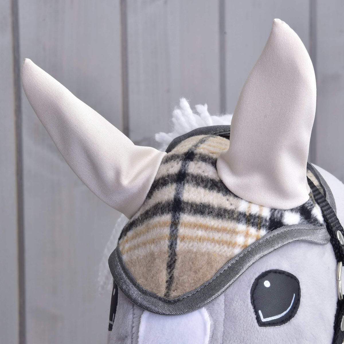 Close-up of the Hobby Horse LarDen Ear Bonnet Cremo England: features soft cream-colored ears, a plaid bridle for fly protection, and a gray mane, shown against a light wooden background.