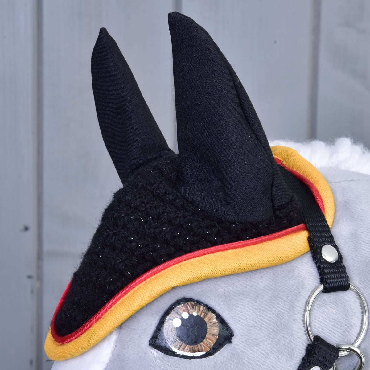 Close-up of a gray hobby horse by Hobby Horse LarDen wearing the "Ear bonnet crocheted Flag" for insect protection, trimmed in yellow and red, with one glassy eye, metal bridle ring, and soft mane against a gray wood background.