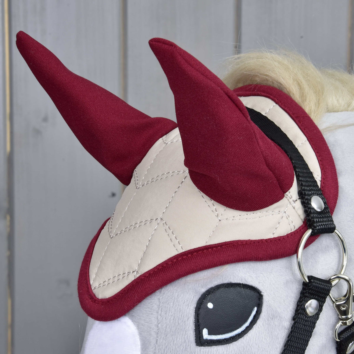 Close-up of a Hobby Horse LarDen with the Ear Bonnet Maroon Star, featuring beige quilting for insect protection, two upright red horn-like shapes, and a black bridle. Light wooden panels are in the background.