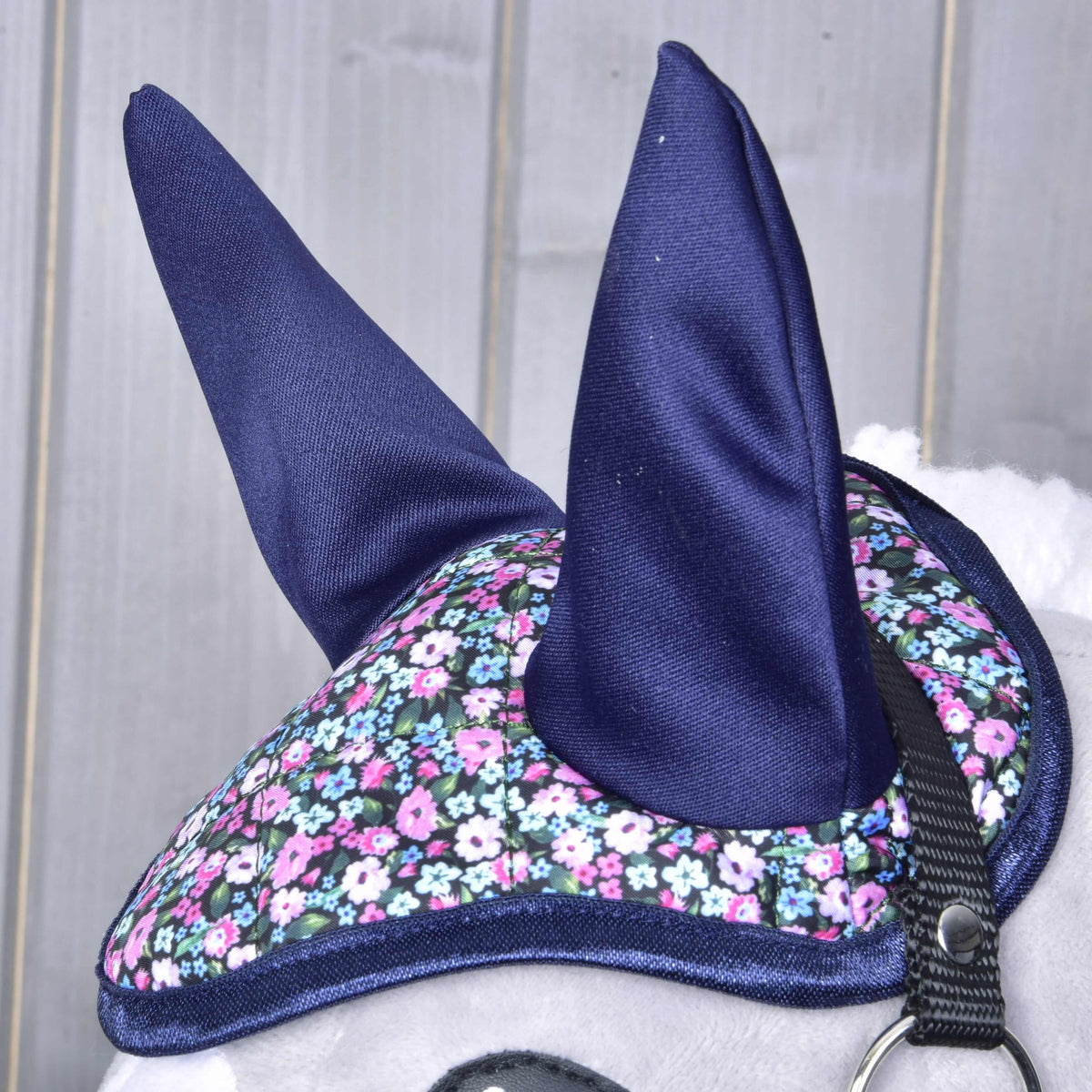 Close-up of a horse wearing the Hobby Horse LarDen Ear Bonnet Flower, featuring dark blue ears and a pink, white, and blue floral pattern; part of the bridle and light gray background show its stylish insect protection.