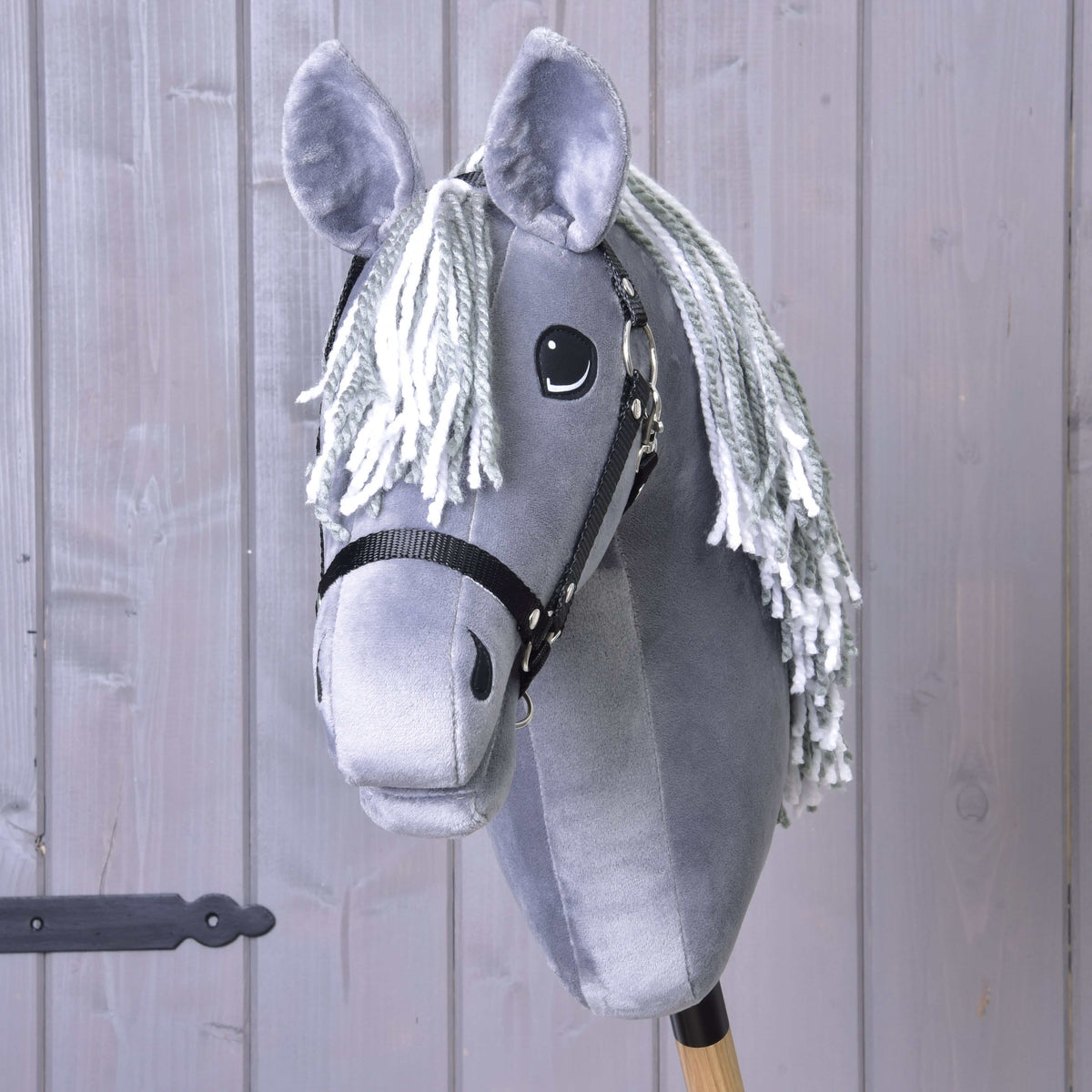The Hobby Horse LarDen Calypso with Black Halter is a gray plush Dutch Warmblood-inspired hobby horse, featuring a white yarn mane and wooden stick handle, shown against a gray wooden wall.