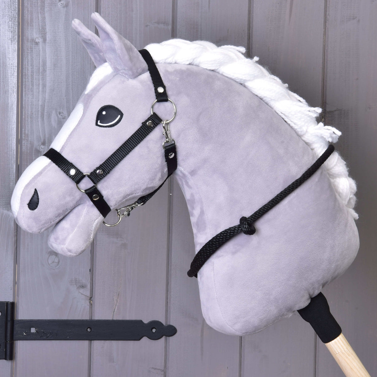 The Cordeo black by Hobby Horse LarDen is a gray plush hobby horse with a white mane on a wooden stick, featuring a black bridle, reins, and handmade cordeo, shown against a gray wooden wall background.