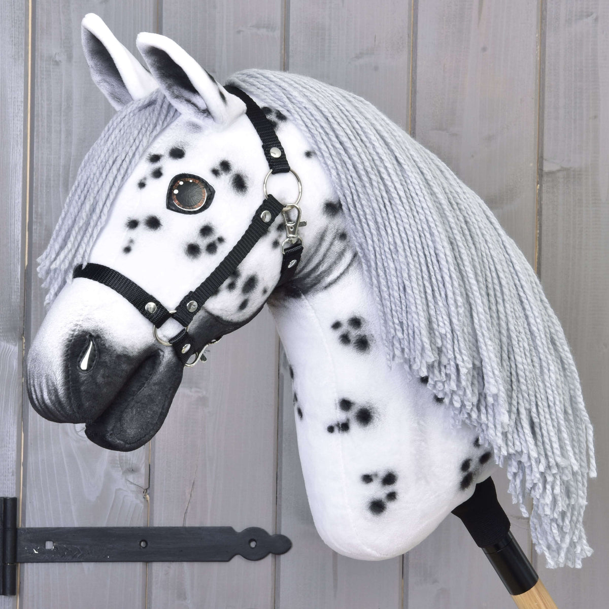 The Hobby Horse LarDen "Chantal with Black Halter" showcases a handcrafted white Knabstrupper-style head with black spots, gray yarn mane, black halter, and wooden stick handle, set against a light wooden plank background.