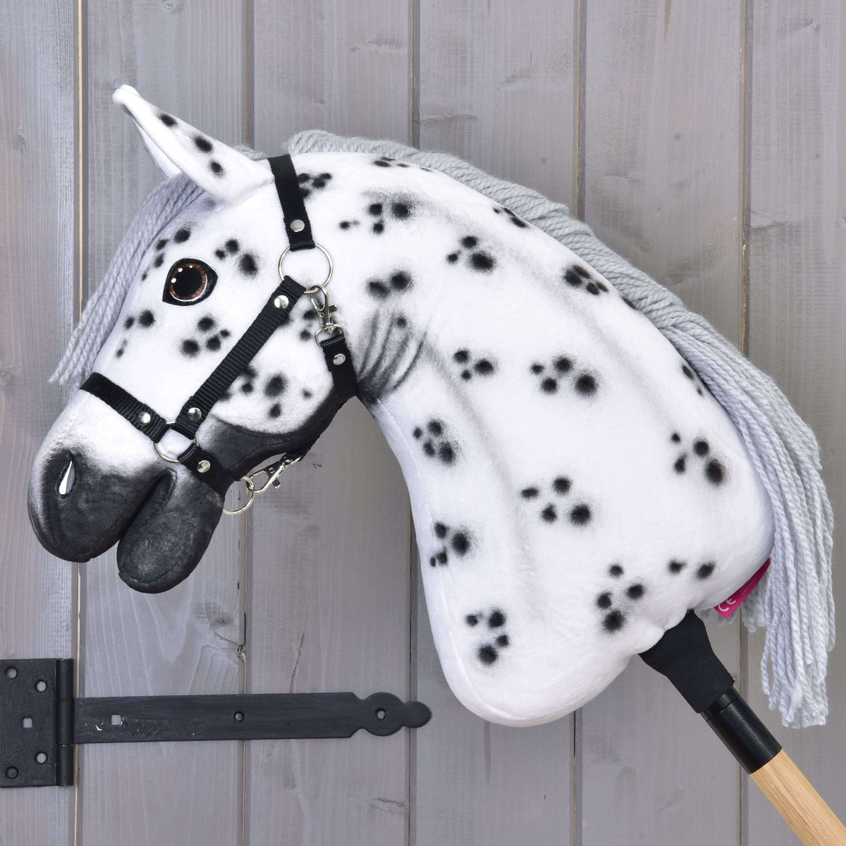 The Hobby Horse LarDen 'Hobby Horse Chantal with Black Halter' features a white coat with black spots, gray yarn mane, black halter, and is mounted on a wooden stick against a light gray wooden wall.