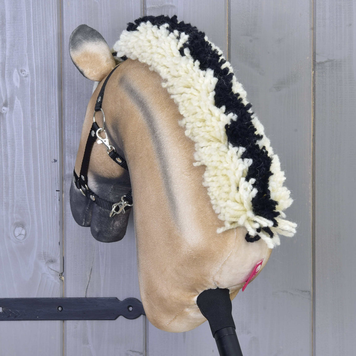 The Hobby Horse LarDen "Colby" features a beige face, black and white yarn mane, black halter, and pink nostrils, mounted on a wooden stick against a gray wooden wall.