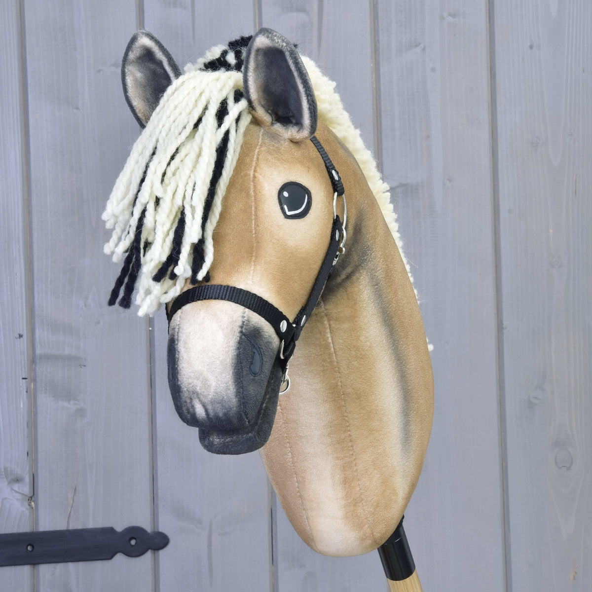 A Hobby Horse LarDen "Colby" with a beige plush head, white and black yarn mane, black halter, and wooden stick stands before a light gray wooden wall.