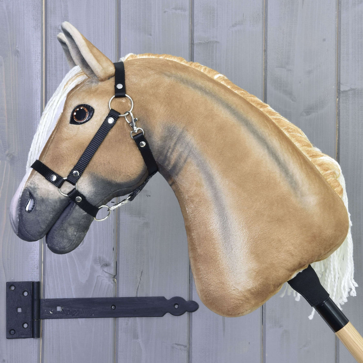 The Hobby Horse LarDen "Hobby Horse Connie with Black Halter" features a plush tan body, white mane, and black halter, shown mounted on a wooden stick against a gray wooden wall.