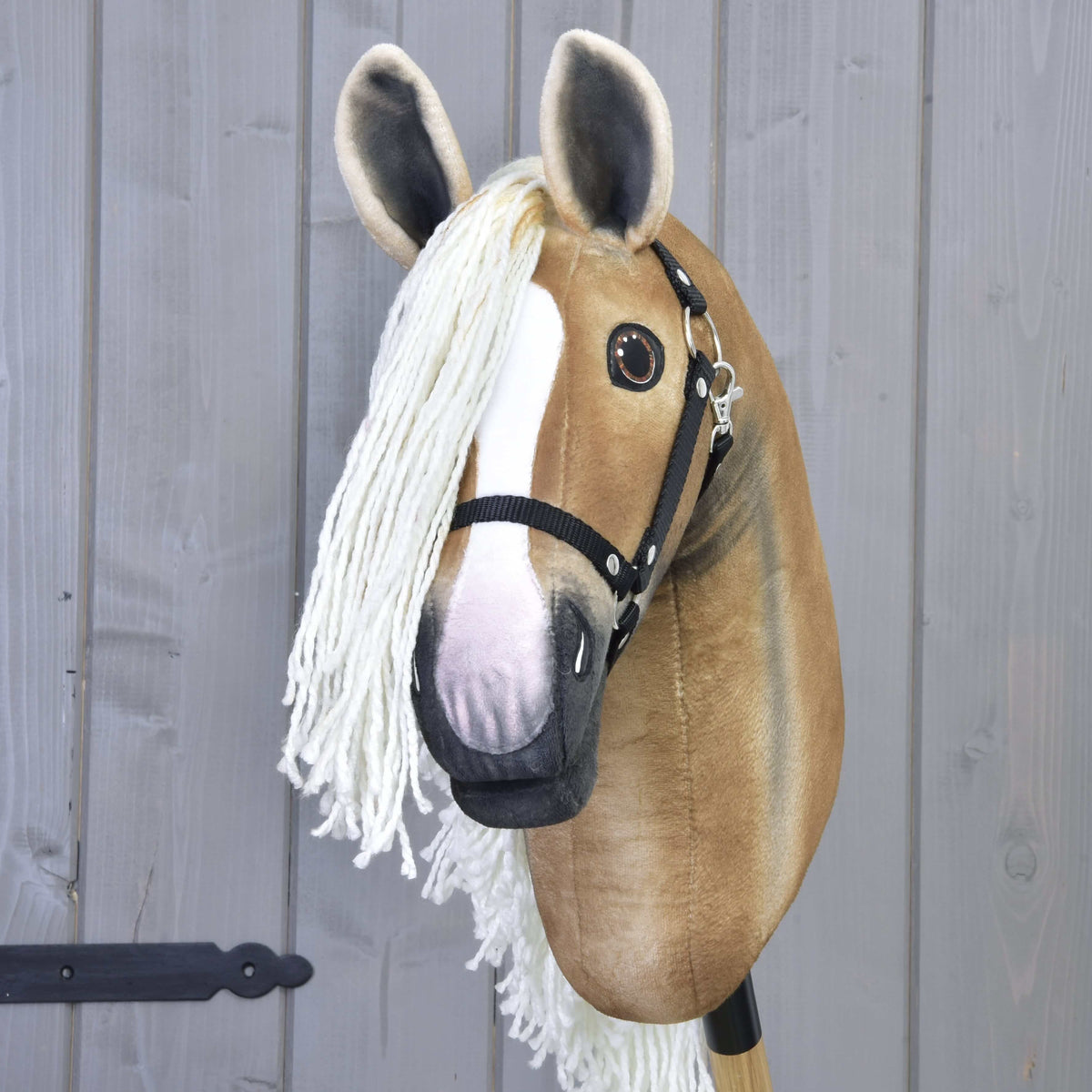 The Hobby Horse LarDen "Hobby Horse Connie with Black Halter" features a handmade tan and white face, large eyes, long white yarn mane, and black halter, mounted on a stick in front of a gray wooden wall.