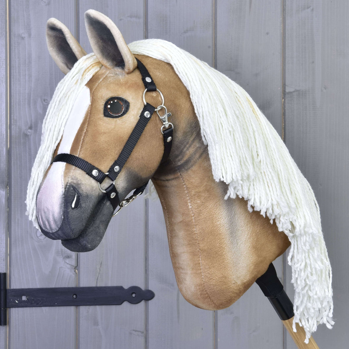 The Hobby Horse LarDen's Hobby Horse Connie with Black Halter features a realistic light tan face, white blaze, black halter, and long white yarn mane on a wooden stick, displayed against a gray wooden wall.