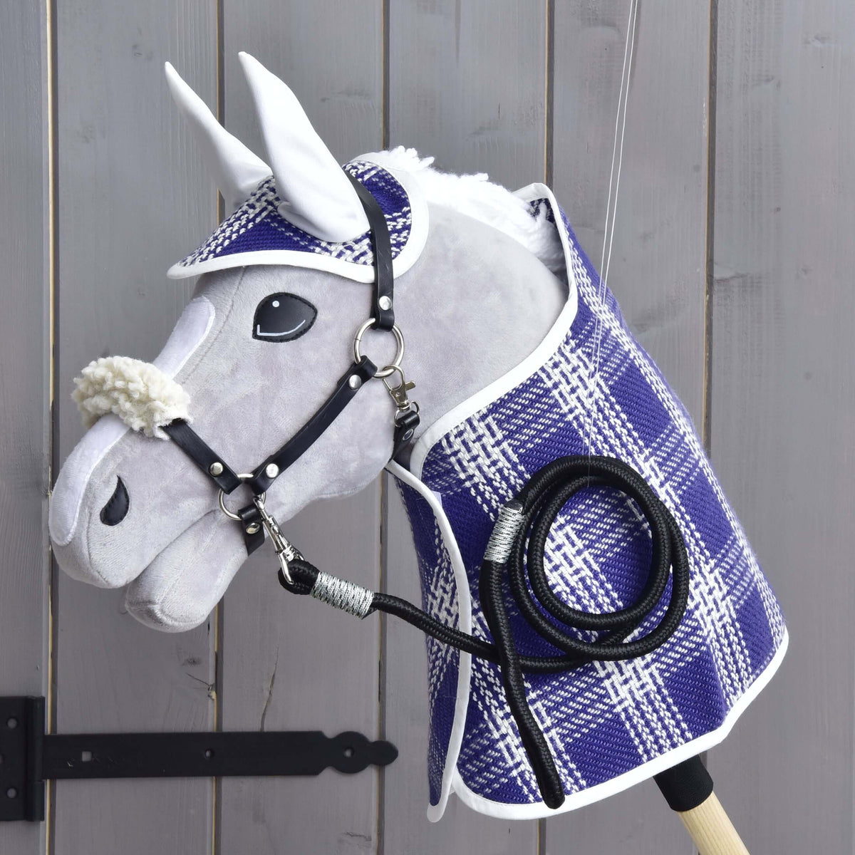 A Hobby Horse LarDen Set Cube, featuring a blanket, ear bonnet, lead rope, and fur halter, is displayed on a wooden wall near a black gate hinge. The accessories highlight the quality and charm of Hobby Horse LarDen products.