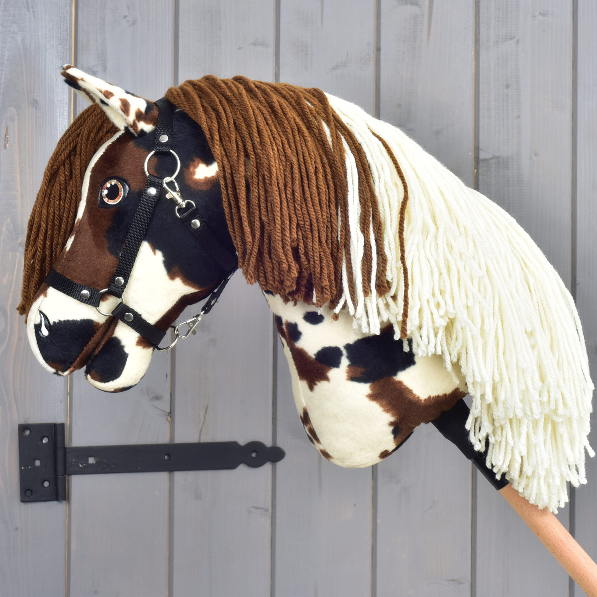 Introducing the Hobby Horse Dakota with Black Halter by Hobby Horse LarDen—a handmade creation inspired by the wild spirit of North America's legendary free-roaming horses. Featuring distinctive brown and white spots, a soft brown yarn mane, a cream yarn forelock, and mounted on a wooden stick, this hobby horse stands proudly against a gray wooden wall as an homage to the untamed Mustang.