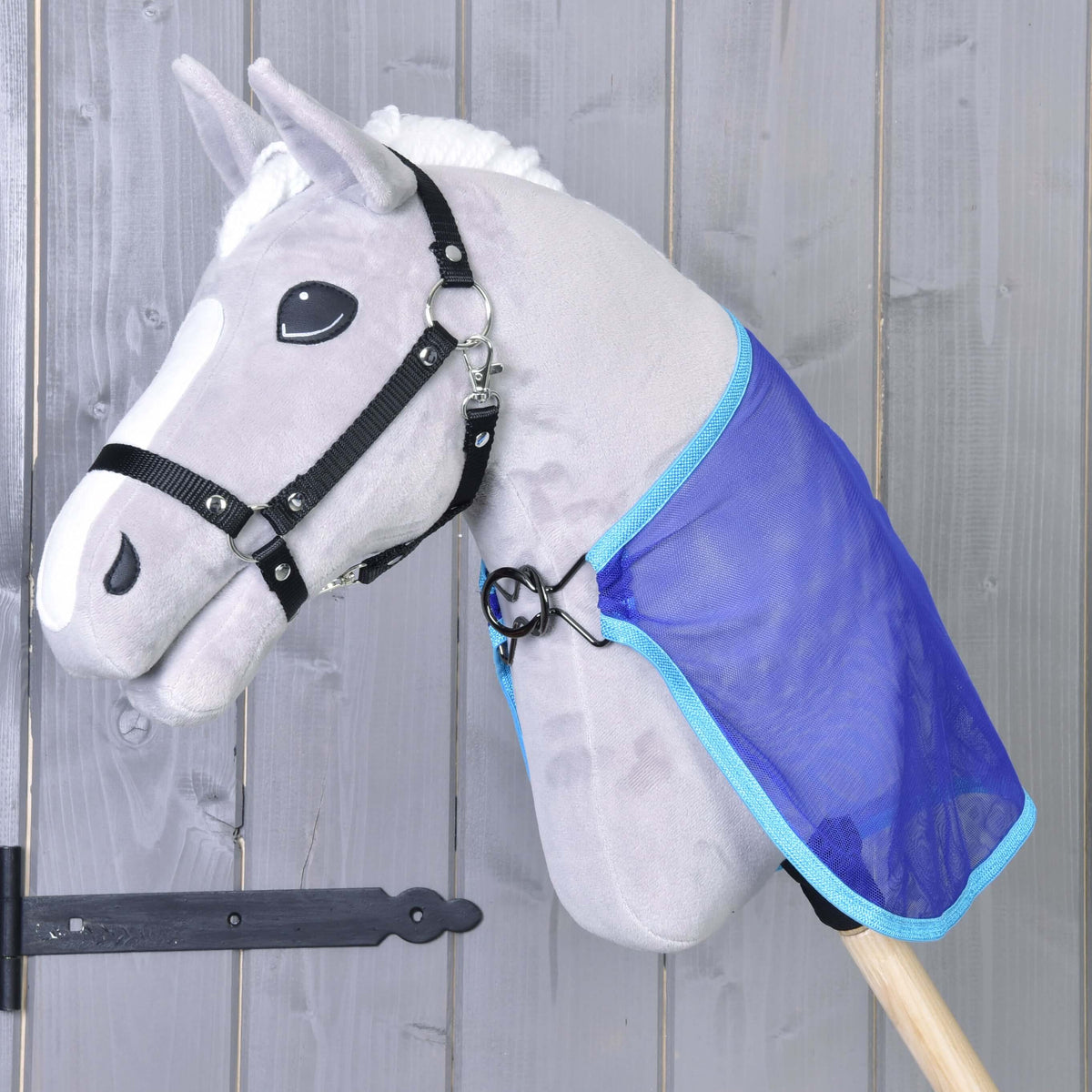 A gray Hobby Horse LarDen toy with a black bridle and white mane wears a blue Fly sheet cape. The horse features cartoon-like eyes and is mounted against a wooden wall.