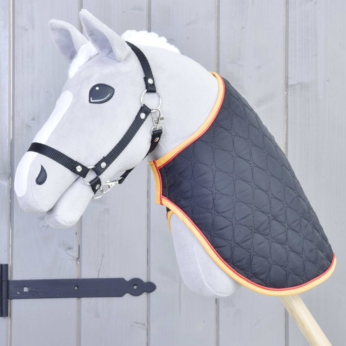 The Hobby Horse LarDen Blanket Flag, a gray hobby horse toy with a black bridle and quilted blanket with orange trim, is mounted on a wooden stick in front of a gray wooden fence.