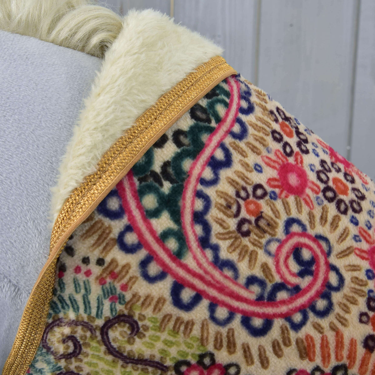 A close-up of Blanket Jungle by Hobby Horse LarDen features vibrant floral and spiral patterns with gold trim and cream faux fur, draped on light gray—ideal as a chic horse training blanket or for stylish protection.