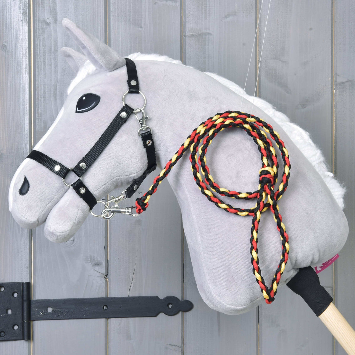 The Hobby Horse LarDen "Hobby Horse - rope stripe Flag" is a gray plush hobby horse with a stick body, white mane, black bridle, and a braided red, yellow, and black leash, shown against a wooden background.