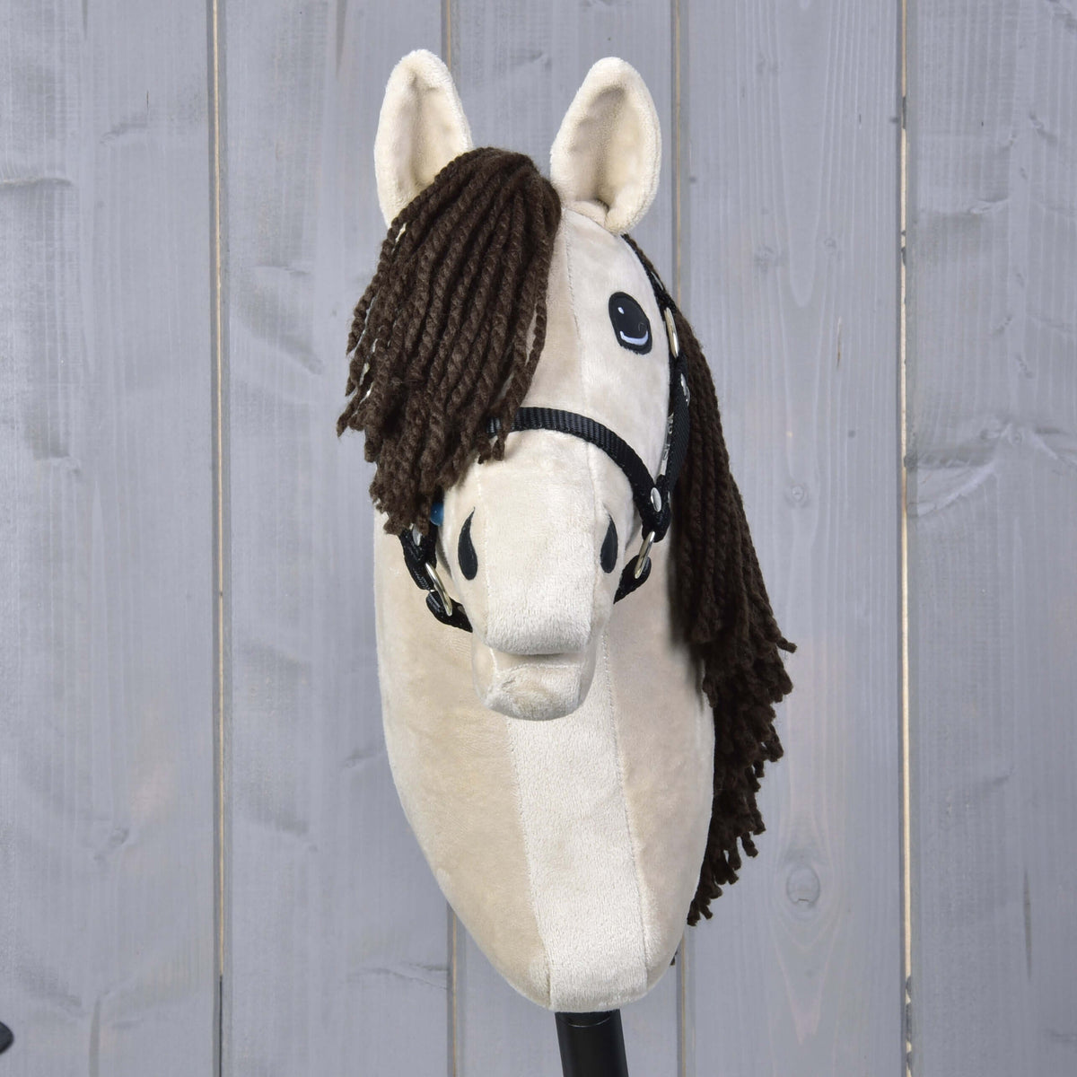 The Hobby Horse LarDen "Hobby Horse Frederik LD with black halter" stands upright in front of a light gray wooden fence, featuring a beige body and brown yarn mane.