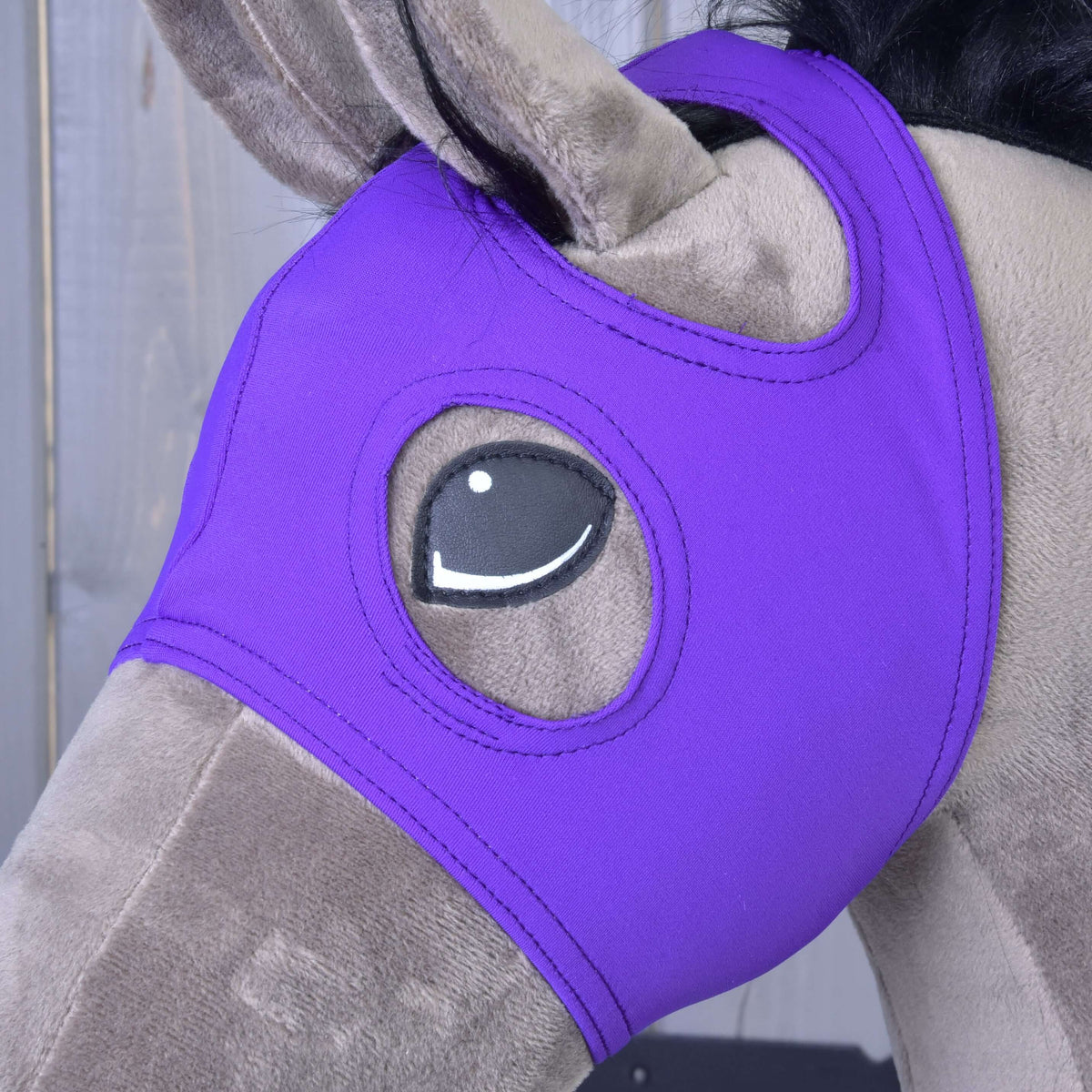 A plush toy horse wears a Hobby Horse LarDen Horse Mask without ears in Indigo, made from washable PES, covering its head with eye openings, set against a wooden background.