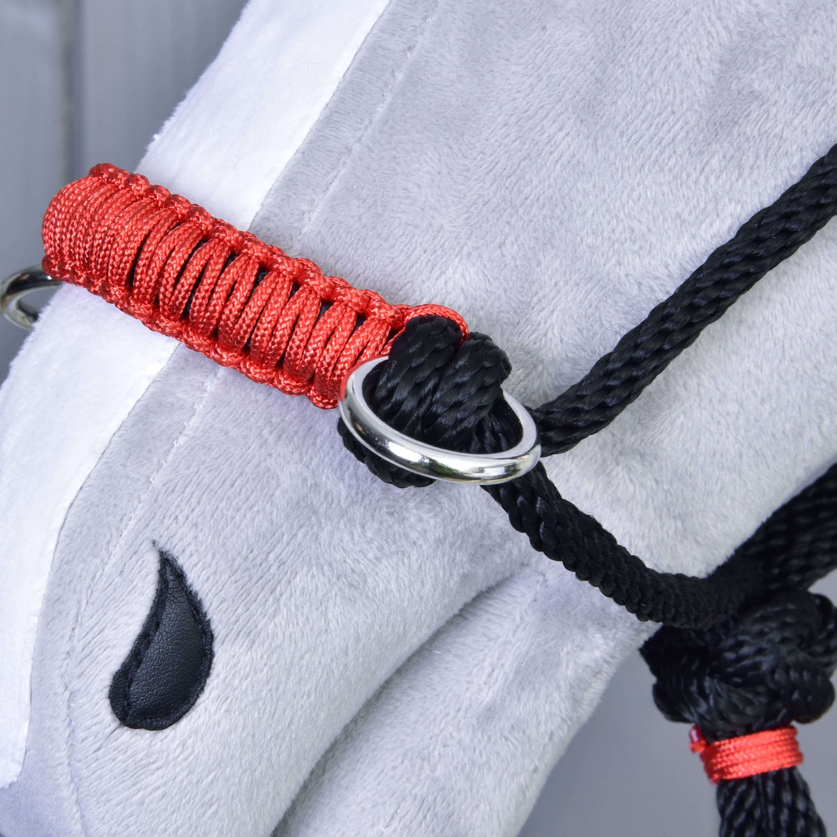 Close-up of a gray stuffed horse’s head wearing the Hobby Horse LarDen Rope halter Spark in red and black with a metal ring, plus a black embroidered teardrop near the eye. Ideal as a unique hobby horse accessory.