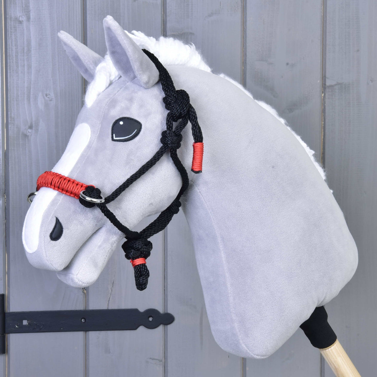 The Hobby Horse LarDen Rope halter Spark is a gray hobby horse toy with a white mane and black-and-red rope halter, mounted on a wooden stick and set in front of a gray wooden wall.