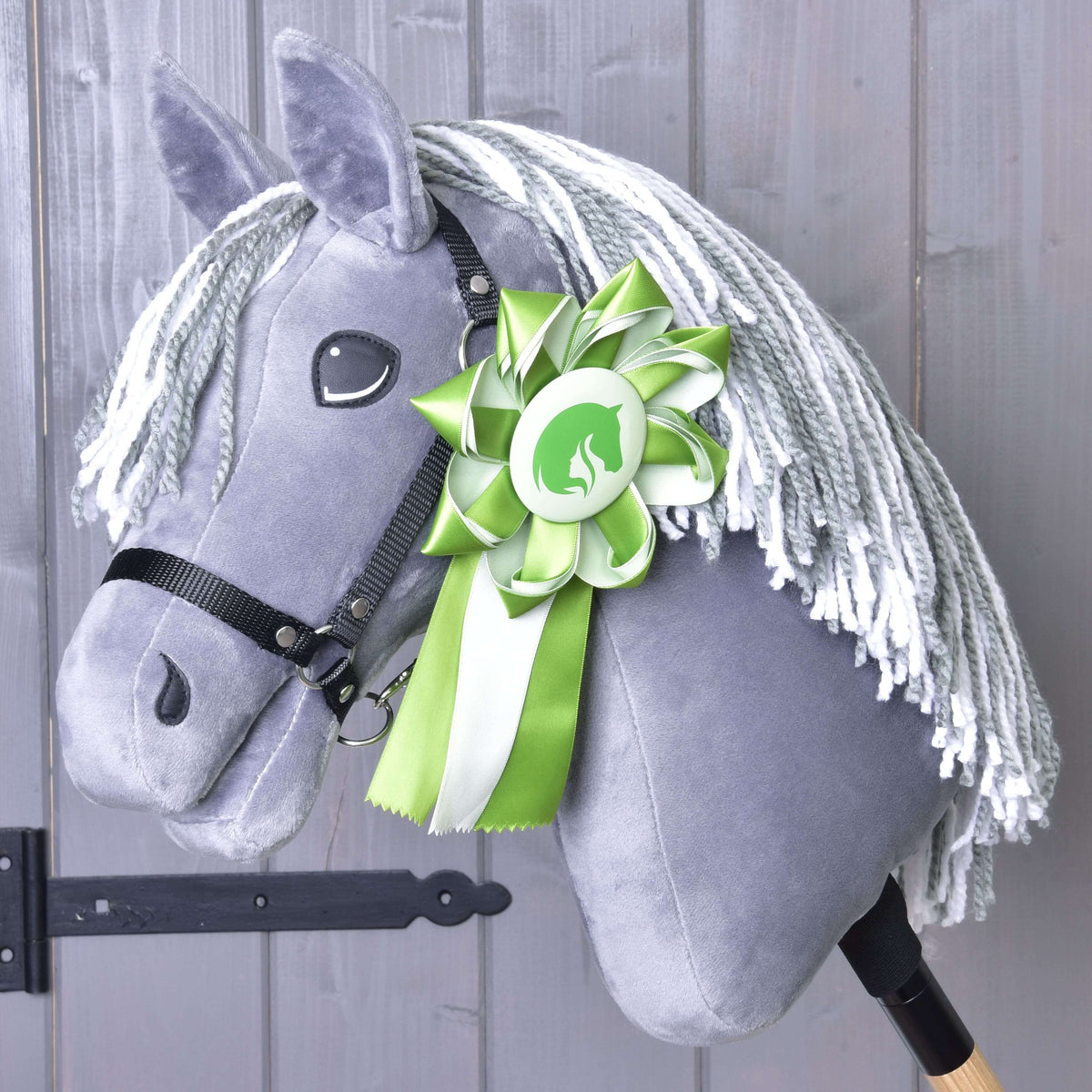 A gray plush Hobby Horse Ribbon Green by Hobby Horse LarDen features a white yarn mane, a black bridle, and a large green and white winning horse ribbon. The wooden stick is visible against a backdrop of wooden paneling.
