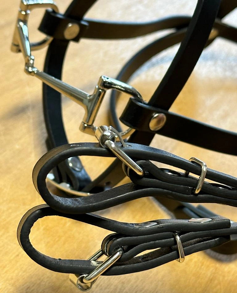 Nærbillede af en sort trense og tøjler med blanke spænder og ringe i sølvfarvet metal på en lysebrun overflade.