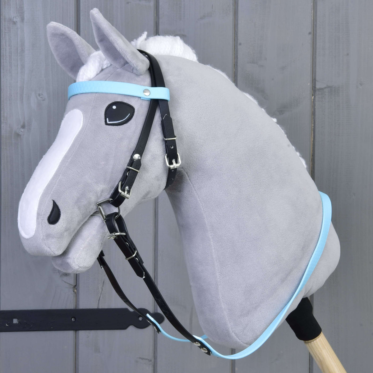 A gray plush hobby horse with a white mane, fitted with the Hobby Horse LarDen Complete bridle without noseband in Light Blue, mounted on a wooden stick and pictured before a gray wooden wall.