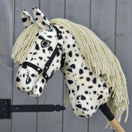 The Hobby Horse LarDen Lord Valentine with Black Halter features a plush spotted coat, long cream-colored mane, button eye, and is mounted on a wooden stick in front of gray wooden panels.