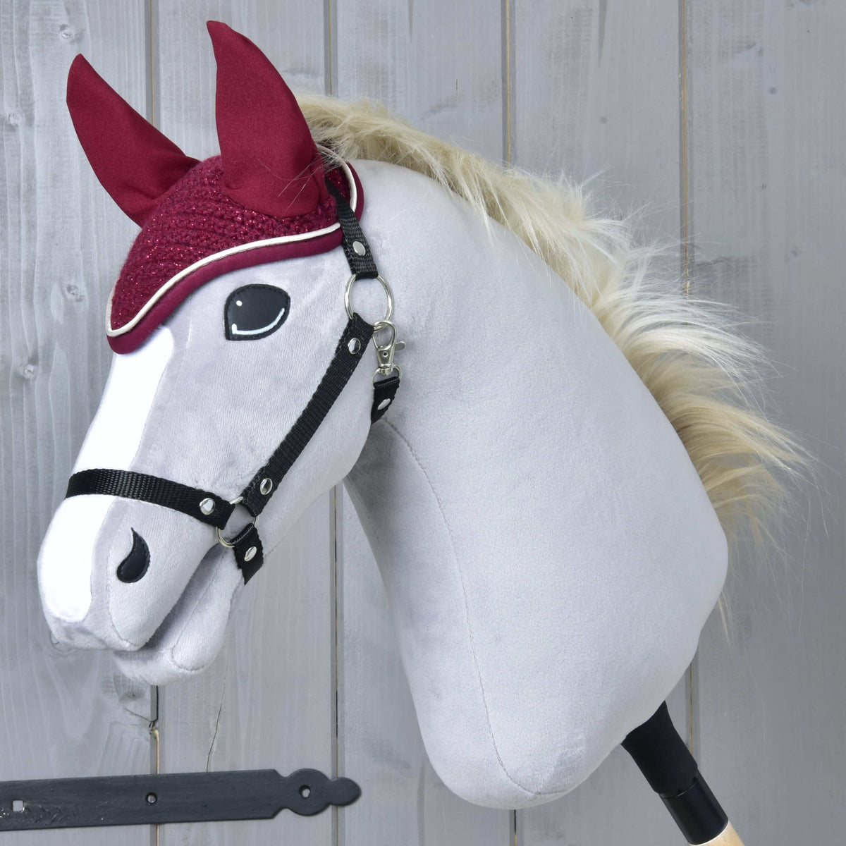 The Hobby Horse LarDen Ear Bonnet Crocheted Lux Maroon is shown on a plush stick horse toy with a white face, black bridle, fluffy blond mane, maroon fabric ears, and matching bonnet against a gray wooden background.