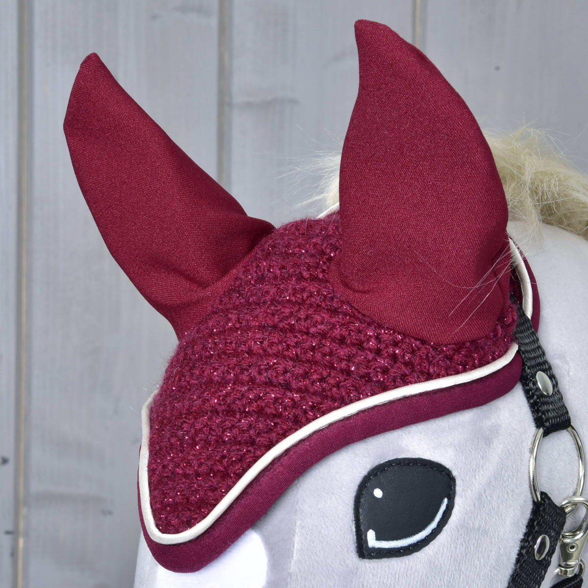 A close-up of the Hobby Horse LarDen Ear Bonnet Crocheted Lux Maroon, featuring tall, pointed ears and white trim on a white hobby horse, set against a wooden background.