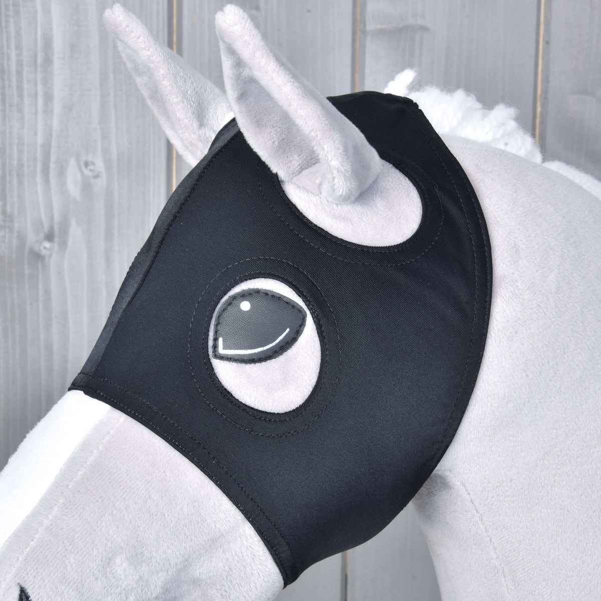 A close-up of a white plush toy horse wearing the Hobby Horse LarDen "Horse Mask without ears Flamingo," featuring PES material with eye cutouts. The background displays wooden panels.