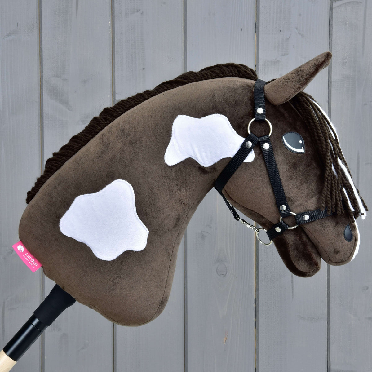 The Hobby Horse LarDen Monte with Black Halter is a brown plush hobby horse with white patches and a black halter, mounted on a wooden stick, shown against a gray wooden plank background.