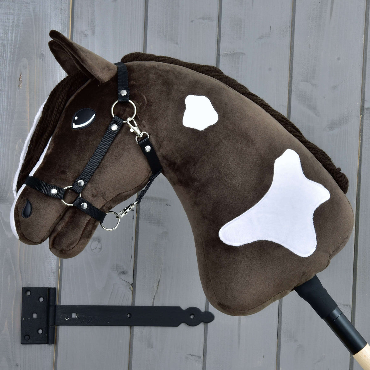 The Hobby Horse LarDen "Hobby Horse Monte with Black Halter" is a brown and white plush hobby horse inspired by the American Paint Horse, featuring a friendly face and white patches, mounted on a wooden gray wall.