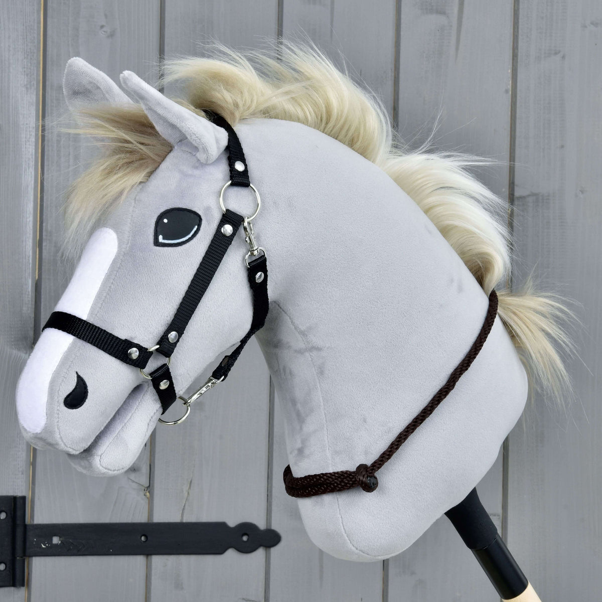 The Cordeo Chocolate by Hobby Horse LarDen is a gray plush hobby horse with a black bridle and cream rope mane on a wooden stick, shown against a gray wooden panel background.