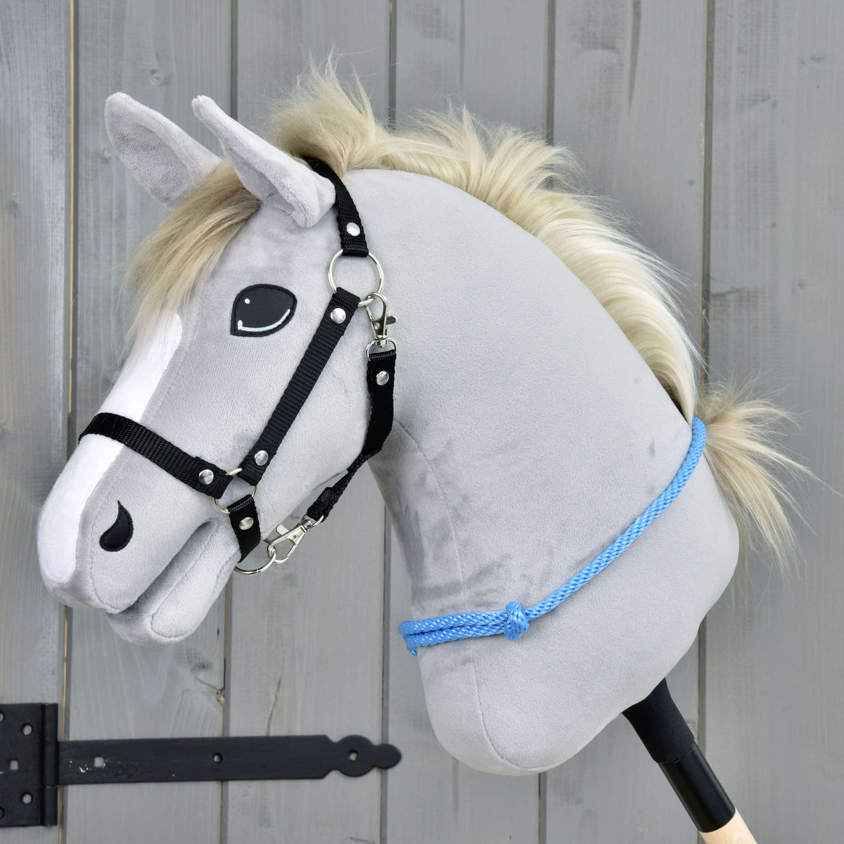 The Hobby Horse LarDen "Cordeo Sky" is a gray plush hobby horse with a white mane, black bridle, and blue rope, designed for riding with or without the bridle. It stands against a wooden background and has a friendly expression.